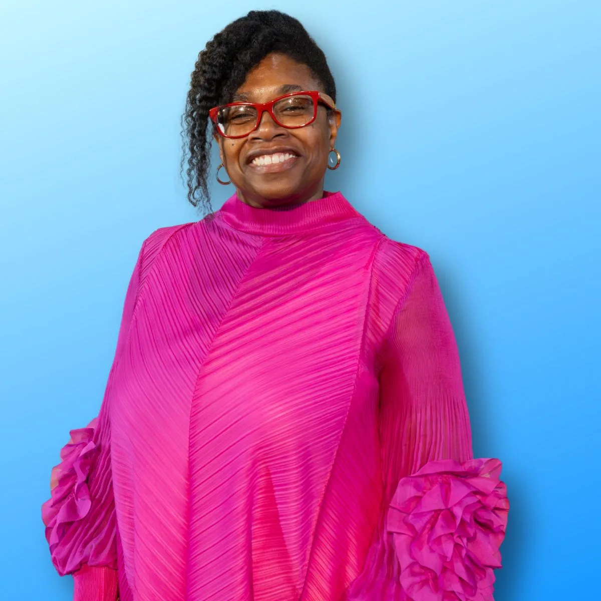 pastorv Smiling woman in a bright pink, textured dress with ruffled sleeves, wearing red glasses, against a blue background.